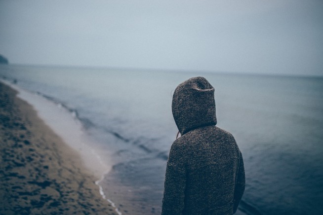 person in grey hoodie looks over sea/beach