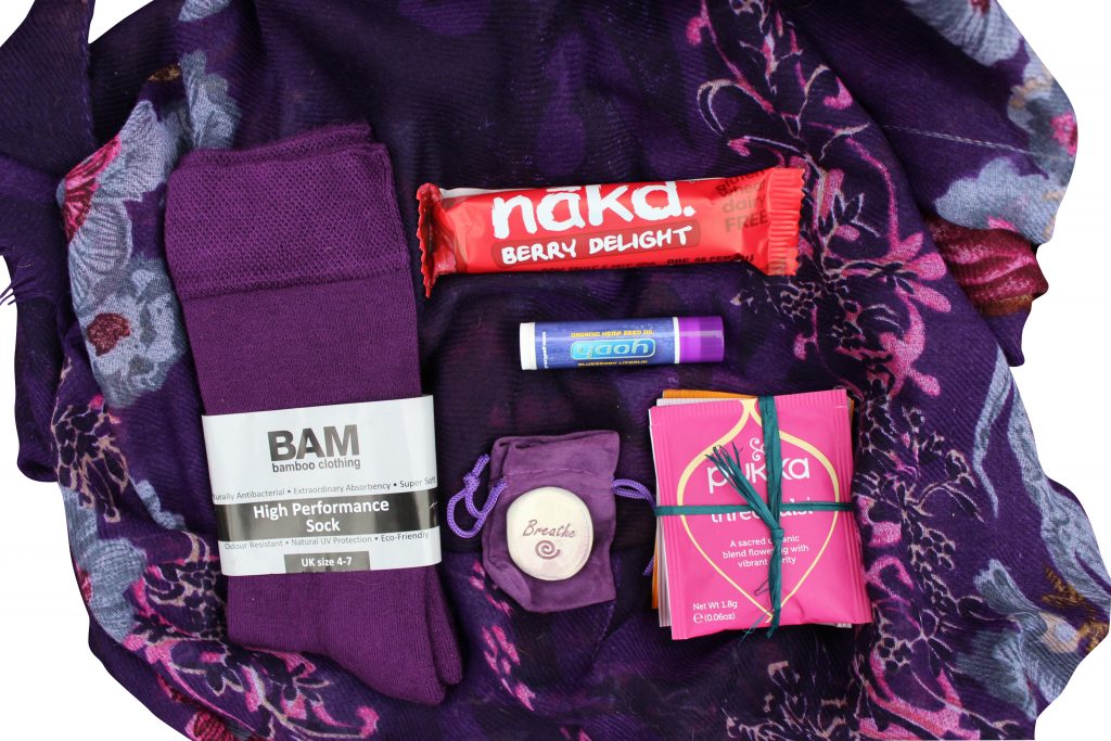 contents of a self care subscription box on a purple cloth shot taken from above. Contains socks, palm stone, tea bags, lip balm and snack ba